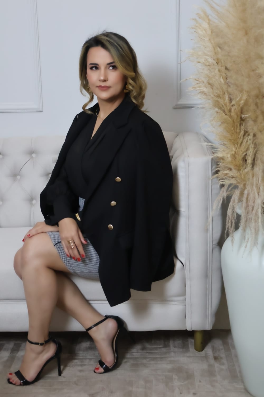 Luana Ribeiro sentada em um sofá claro, com blazer preto, em ambiente de escritório.