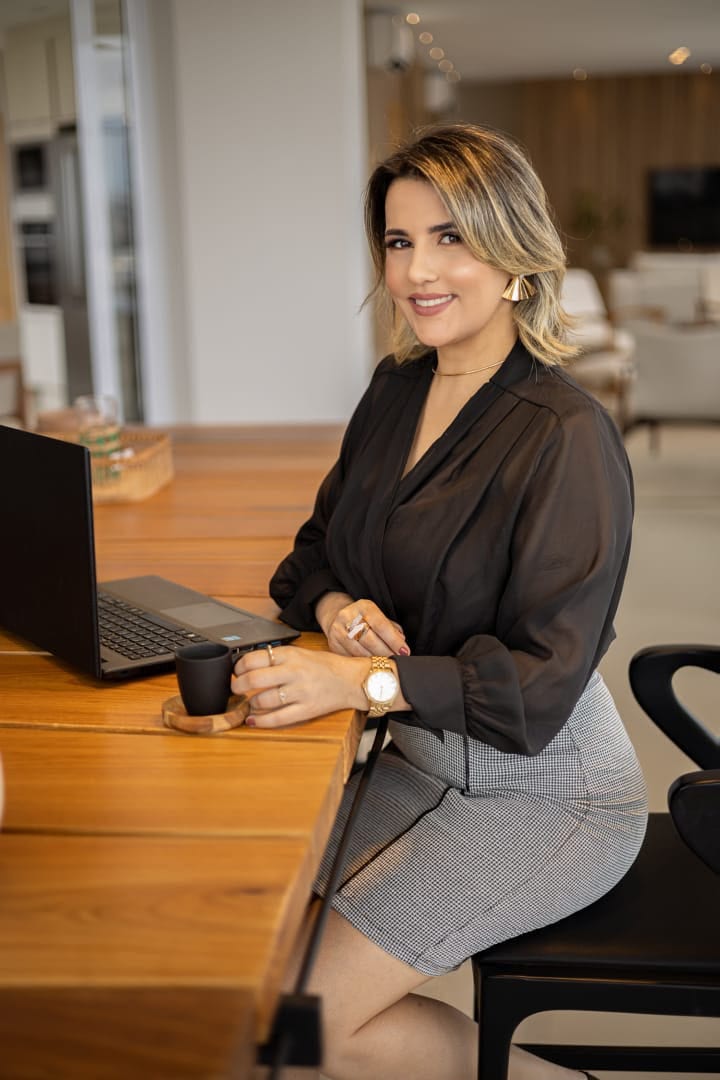 Luana Ribeiro sorrindo, sentada a uma mesa de madeira com notebook e uma xícara de café.