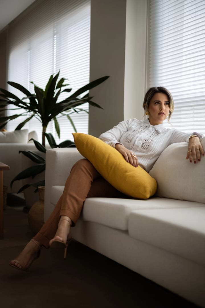 Luana Ribeiro sentada em um sofá claro, com blusa branca, ao lado de uma almofada amarela e plantas, em ambiente iluminado por luz natural.